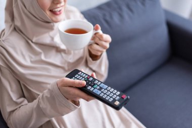 Bir fincan çay ve uzaktan kumandayla evde televizyon izleyen gülümseyen Müslüman kadın manzarası.