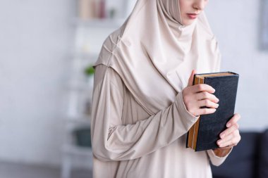 partial view of arabian woman in hijab holding koran while praying at home clipart