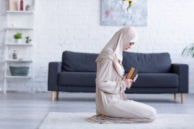 side view of muslim woman praying with koran on floor at home clipart