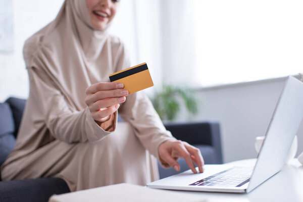 cropped view of blurred muslim woman holding credit card near laptop