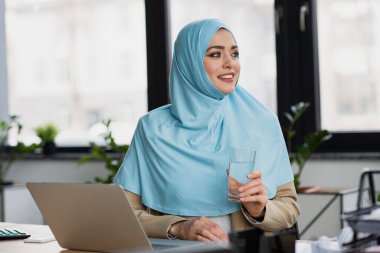 Bir bardak suyla başarılı Müslüman iş kadını ofisteki dizüstü bilgisayarın yanına bakıyor.