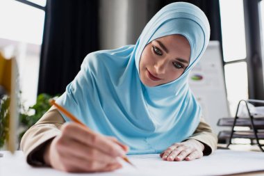 Genç Müslüman tasarımcı ön planda bulanık kalemle çizim yapıyor.