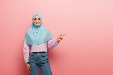 joyful arabian woman in jeans and hijab pointing aside with finger on pink background clipart