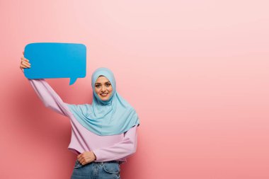 Mavi tesettürlü neşeli Arap kadın pembe arka planda konuşma balonu tutuyor.