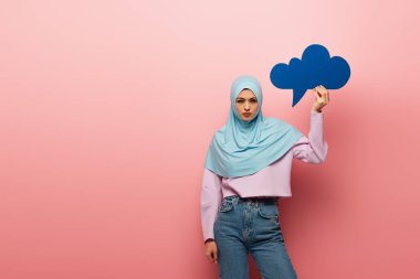 Müslüman kadın tesettürlü ve kot pantolonlu pembe arka planda mavi düşünce balonu tutuyor
