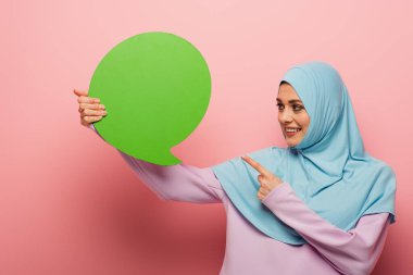 smiling muslim woman pointing at green thought bubble on pink background clipart