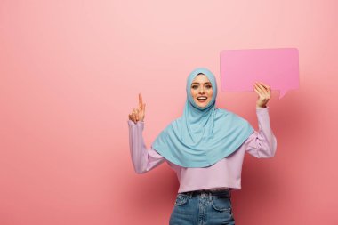 positive muslim woman pointing up with finger while holding speech bubble on pink background clipart