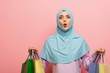 Şaşkın müslüman kadın elinde pembe arka plandaki alışverişleri tutarken kameraya bakıyor.