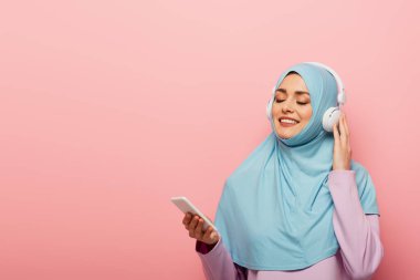 Akıllı telefonlu neşeli Müslüman kadın pembe kulaklıkla müzik dinliyor.