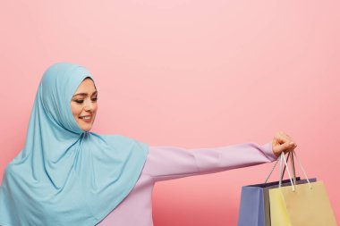 Mavi tesettürlü neşeli Arap kadın pembe arka planda çok renkli alışveriş torbaları gösteriyor.