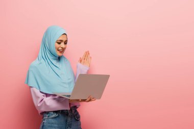 Neşeli Müslüman kadın pembe arka planda video görüşmesinde el sallıyor.