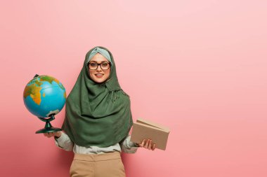 Dünya ve ders kitaplarıyla neşeli Müslüman öğretmen pembe arka planda kameraya bakıyor.