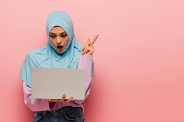 surprised muslim woman pointing with finger while looking at laptop on pink background clipart