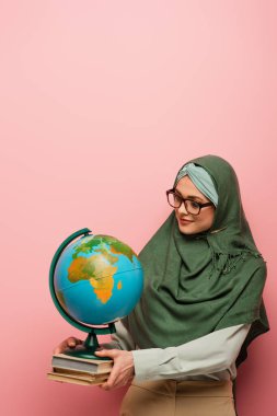 Yeşil tesettürlü Arapça öğretmeni pembe arka planda kitap ve dünya tutuyor.
