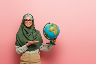 smiling muslim teacher in green hijab pointing at globe on pink background clipart
