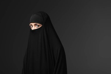 young muslim woman in black traditional clothing looking at camera isolated on black clipart