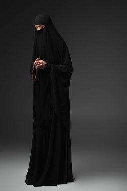 Siyah takkeli Müslüman kadının koyu gri arka planda tespihle dua edişinin tam boy görüntüsü.