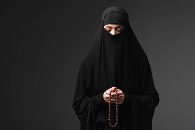 muslim woman in black niqab praying with rosary isolated on black clipart