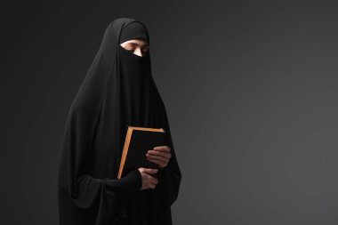 young muslim nun holding koran while praying with closed eyes isolated on black clipart