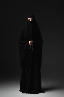 full length view of muslim nun in black niqab and abaya praying with rosary on black background clipart