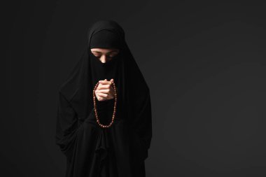 muslim nun in black traditional clothing praying with rosary isolated on black clipart