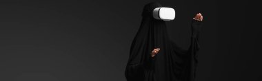 muslim nun gesturing while gaming in vr headset isolated on black, banner clipart