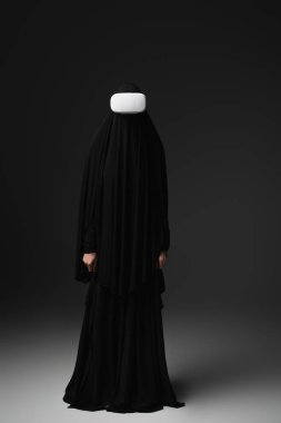 full length view of muslim nun in black abaya and vr headset on black background clipart