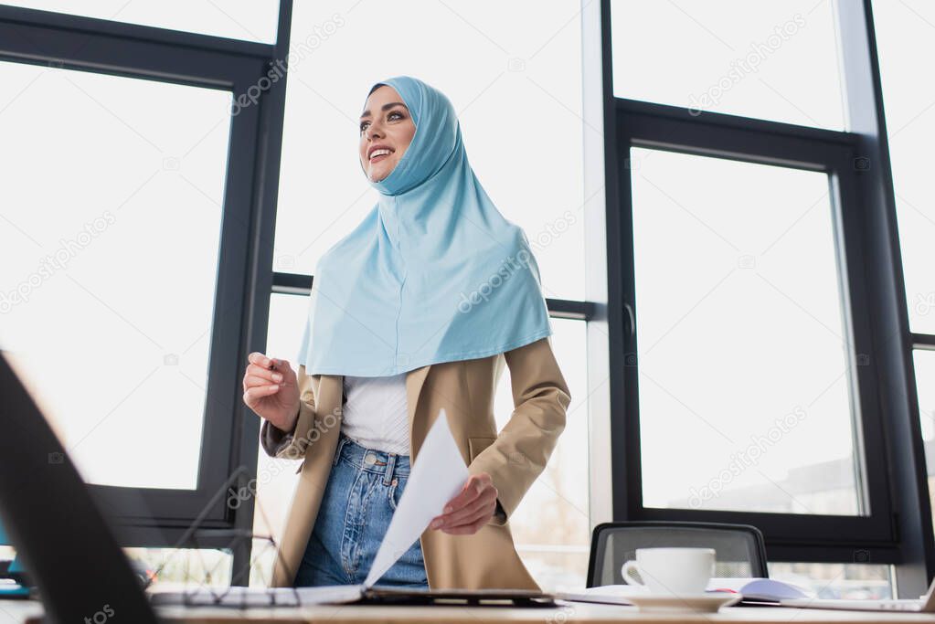 smiling muslim businesswoman holding paper while standing against window in office