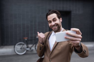 Bulanık ve mutlu genç iş adamı dışarıda cep telefonuyla selfie çekiyor.