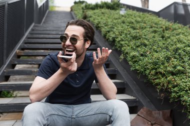 Güneş gözlüklü ve polo tişörtlü mutlu adam dışarıda cep telefonundan sesli mesaj kaydediyor. 