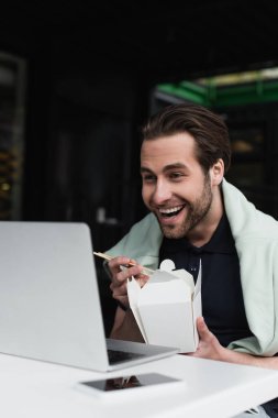 Polo tişörtlü ve kazaklı mutlu bir adam yemek çubukları ve karton kutu tutarken bilgisayarın yanındaki bulanık akıllı telefona bakıyor. 