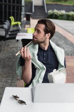 bearded man in polo shirt and sweatshirt holding chopsticks and carboard box near laptop and blurred sunglasses on table clipart