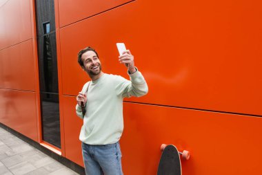 Sweatshirt 'lü neşeli adam turuncu duvarın ve uzun tahtanın yanındaki akıllı telefondan selfie çekiyor. 