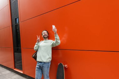Sweatshirt 'lü neşeli adam akıllı telefondan selfie çekiyor ve turuncu duvarın ve uzun panonun yanında barış işareti gösteriyor. 