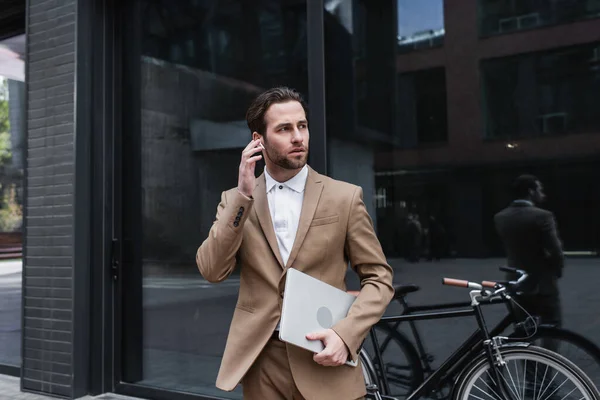 bearded businessman adjusting earphone and holding laptop near building and bike