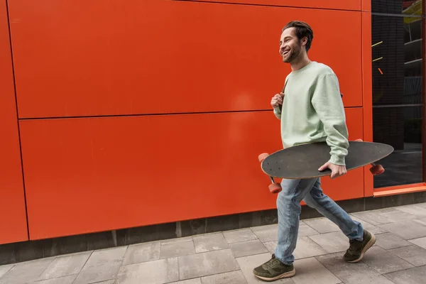 Homem Sorridente Camisola Segurando Longboard Enquanto Caminhava Perto Parede Laranja — Fotografia de Stock