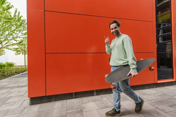 Hombre Feliz Sudadera Sosteniendo Longboard Señalando Con Dedo Mientras Mira — Foto de Stock