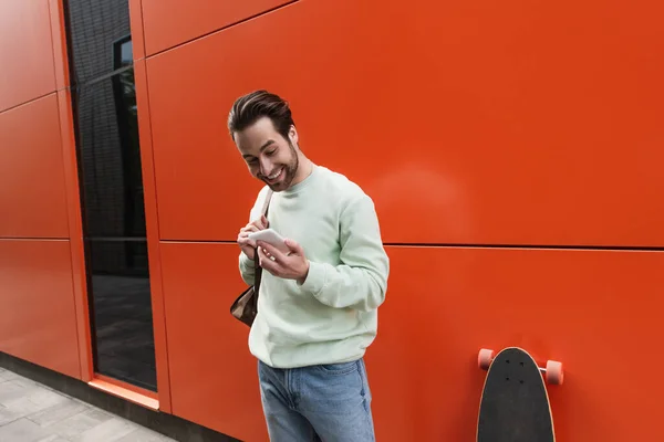 Cheerful Man Sweatshirt Holding Strap Leather Backpack Looking Smartphone Orange — Stock Photo, Image