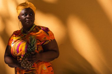 Orta yaşlı Afro-Amerikalı kadın sarı sarıklı ve turuncu üzerinde ananas tutan renkli elbiseli.