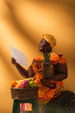 Orta yaşlı Afrikalı Amerikalı kadın meyvelerin yanında oturuyor ve elinde ananas ve vantilatör tutuyor.