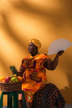 Orta yaşlı gülümseyen Afrikalı Amerikalı kadın meyvelerin yanında oturuyor ve elinde ananas ve vantilatör tutuyor.