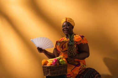 Pozitif orta yaşlı Afro-Amerikalı kadın meyvelerin yanında oturuyor ve elinde ananas ve vantilatör tutuyor.
