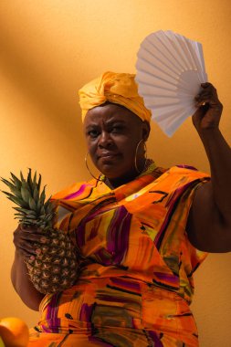 Ciddi orta yaşlı Afro-Amerikalı kadın ananas tutuyor ve turuncu yelpazeyle el sallıyor.