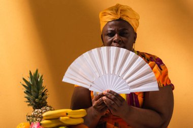 Ciddi orta yaşlı Afrikalı Amerikalı kadın meyvelerin yanında oturuyor ve elinde turuncu yelpaze tutuyor.