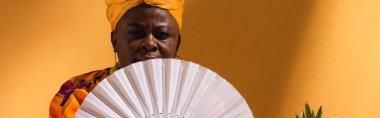 serious middle aged african american woman covering face with fan on orange, banner clipart