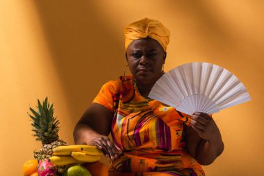 Ciddi bir orta yaşlı Afro-Amerikalı kadın hayranlarıyla turuncu renkli meyvelerin yanında oturuyor.