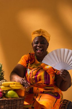 Orta yaşlı gülümseyen Afro-Amerikalı kadın elinde vantilatörle turuncu meyvelerin yanında oturuyor