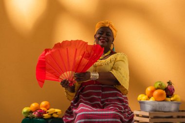 Orta yaşlı gülümseyen Afrikalı Amerikalı kadın meyvelerin yanında oturuyor ve elinde turuncu yelpaze tutuyordu.