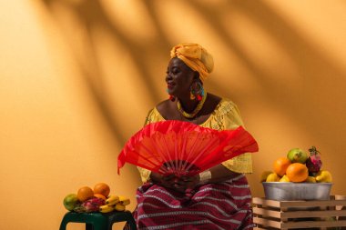 Neşeli orta yaşlı Afrikalı Amerikalı kadın meyve satıyor ve elinde turuncu yelpaze tutuyor.