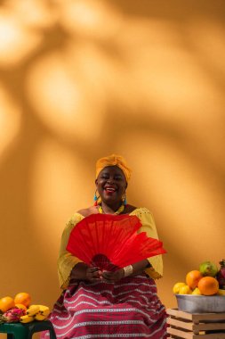 Pozitif orta yaşlı Afrikalı Amerikalı kadın meyve satıyor ve elinde turuncu yelpaze tutuyor.
